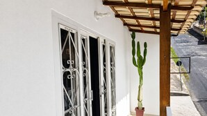 Un edificio blanco con una ventana que tiene barrotes decorativos de metal, un cactus en maceta y una estructura de pérgola de madera.
