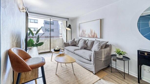 A modern living room with a sofa, a coffee table, a chair, a side table with a plant, and a painting on the wall. - Prime Apartment - MUT y Costanera Center (Las Condes)