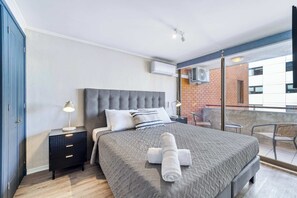 A modern hotel room with a large bed, a bedside table with a lamp, a small table with a chair, and a balcony with a view of a brick building. - Prime Apartment - MUT y Costanera Center (Las Condes)