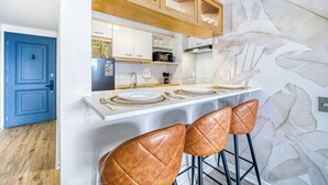 A modern kitchen with white cabinets, a wooden countertop, and a stone wall. There are two brown leather bar stools and a white dining table set with plates and cutlery. - Prime Apartment - MUT y Costanera Center (Las Condes)