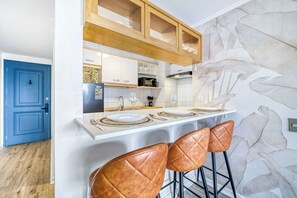 A modern kitchen with white cabinets, a wooden countertop, and a stone wall. There are two brown leather bar stools and a white dining table set with plates and cutlery. - Prime Apartment - MUT y Costanera Center (Las Condes)