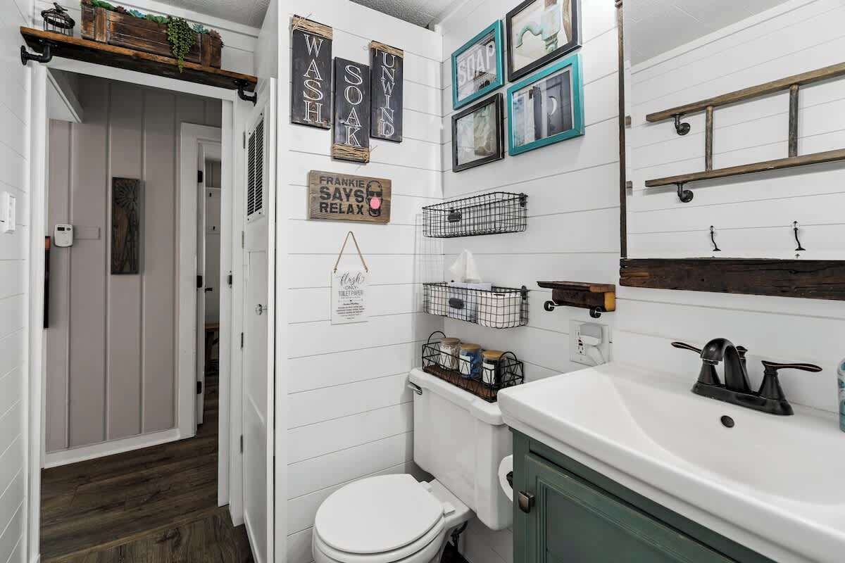Une salle de bain avec des toilettes blanches, un lavabo doté d’un robinet noir et divers éléments de décoration murale, notamment des tableaux encadrés et des panneaux.