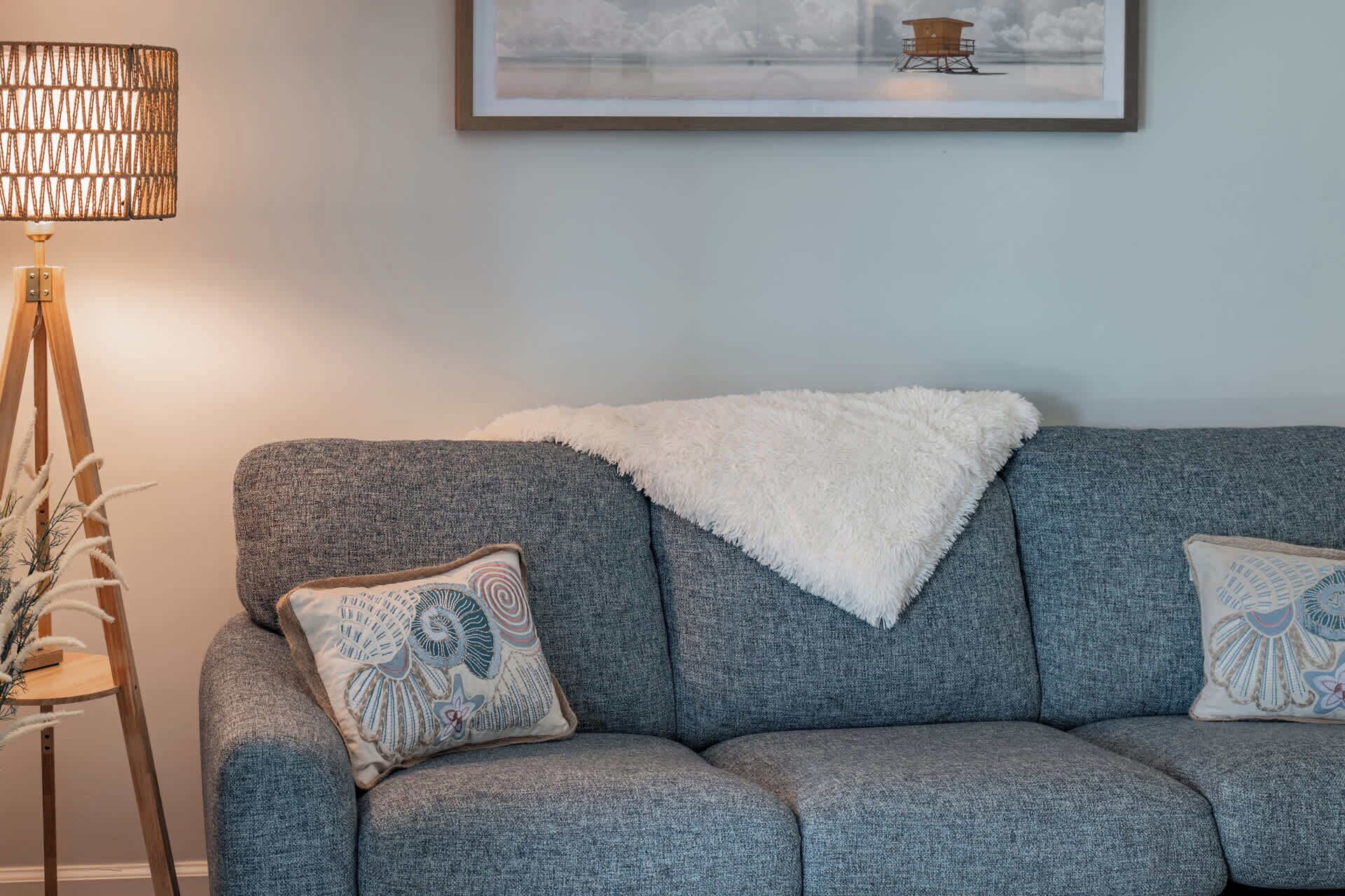 A grey sofa with a white throw blanket and decorative pillows, a floor lamp, and a framed picture on the wall.