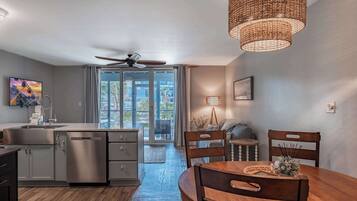 A modern kitchen with a dishwasher, a dining table with chairs, a ceiling fan, and a large window with curtains.