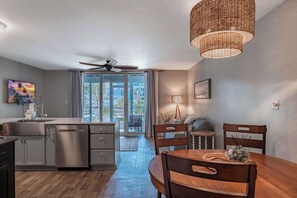 A modern kitchen with a dishwasher, a dining table with chairs, a ceiling fan, and a large window with curtains. - Coastal Calm at Deerfield! Golf Colony 18C (Myrtle Beach)