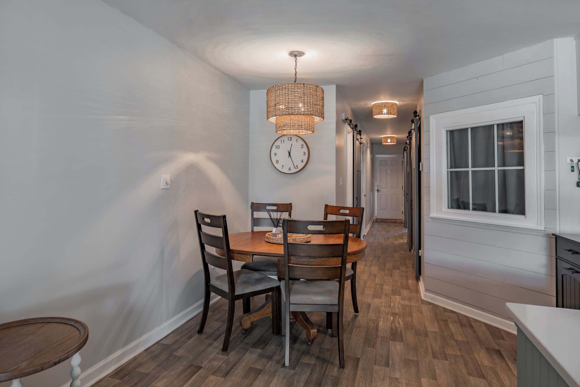 A dining area with a wooden table and chairs, a wall clock, and a hanging light fixture.
