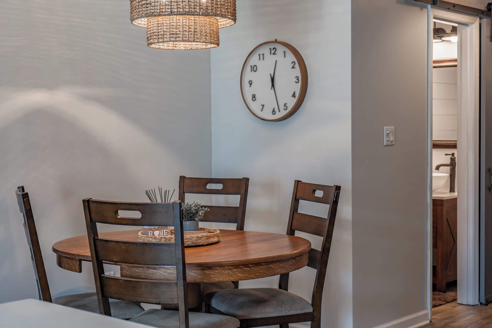A dining area with a round wooden table, four chairs, a wall clock, and a hanging light fixture.