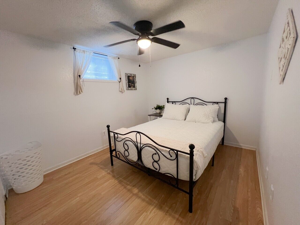 Une chambre à coucher avec un lit, un ventilateur de plafond, une fenêtre avec des rideaux et une petite table de chevet sur laquelle est posée une plante.