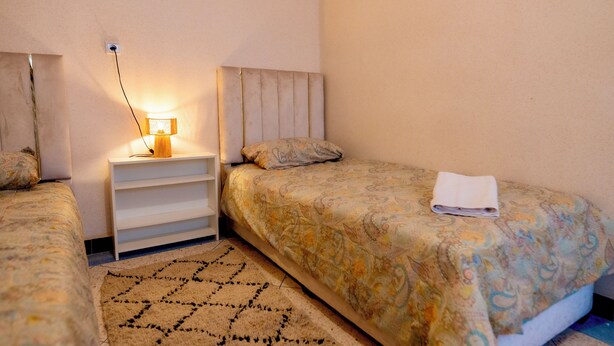 Een slaapkamer met twee bedden, een nachtkastje met een lamp, een kamerkleed met een patroon, en een opgevouwen handdoek op een van de bedden.
