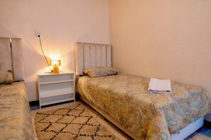 A bedroom with two beds, a bedside table with a lamp, a patterned rug, and a folded towel on one bed. - Bright Apartment with Sunny Rooftop Terrace. (Essaouira)