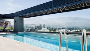 A rooftop pool with clear blue water, a metal ladder, and a view of a cityscape and mountains in the background.
