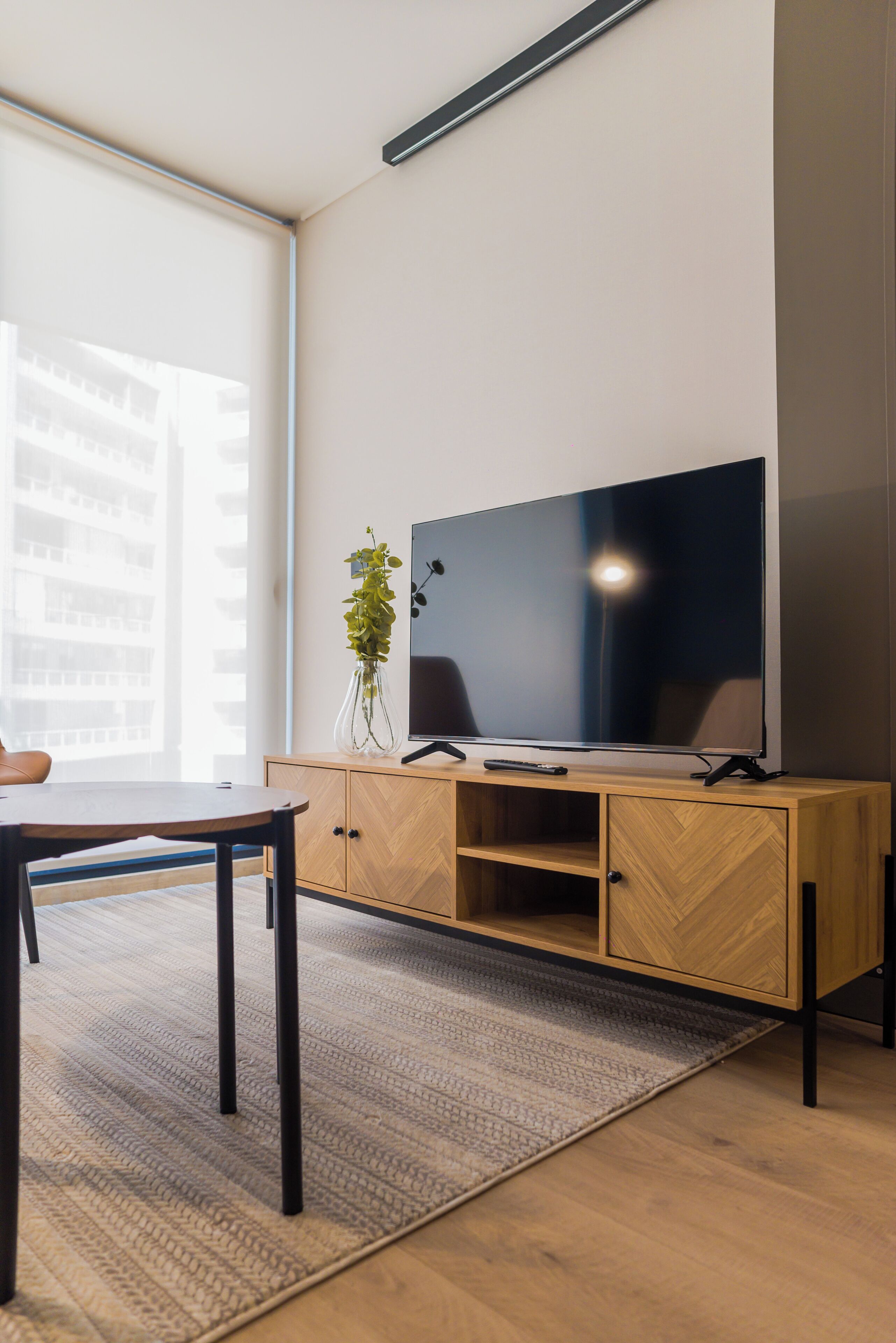 A modern living room with a flat-screen TV on a wooden cabinet, a dining table, and a large window with a view of buildings.