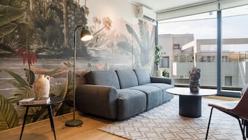 A modern living room with a grey sofa, a round coffee table, and a floor lamp. A tropical mural decorates the wall, and there's a large window offering a view of the outside.