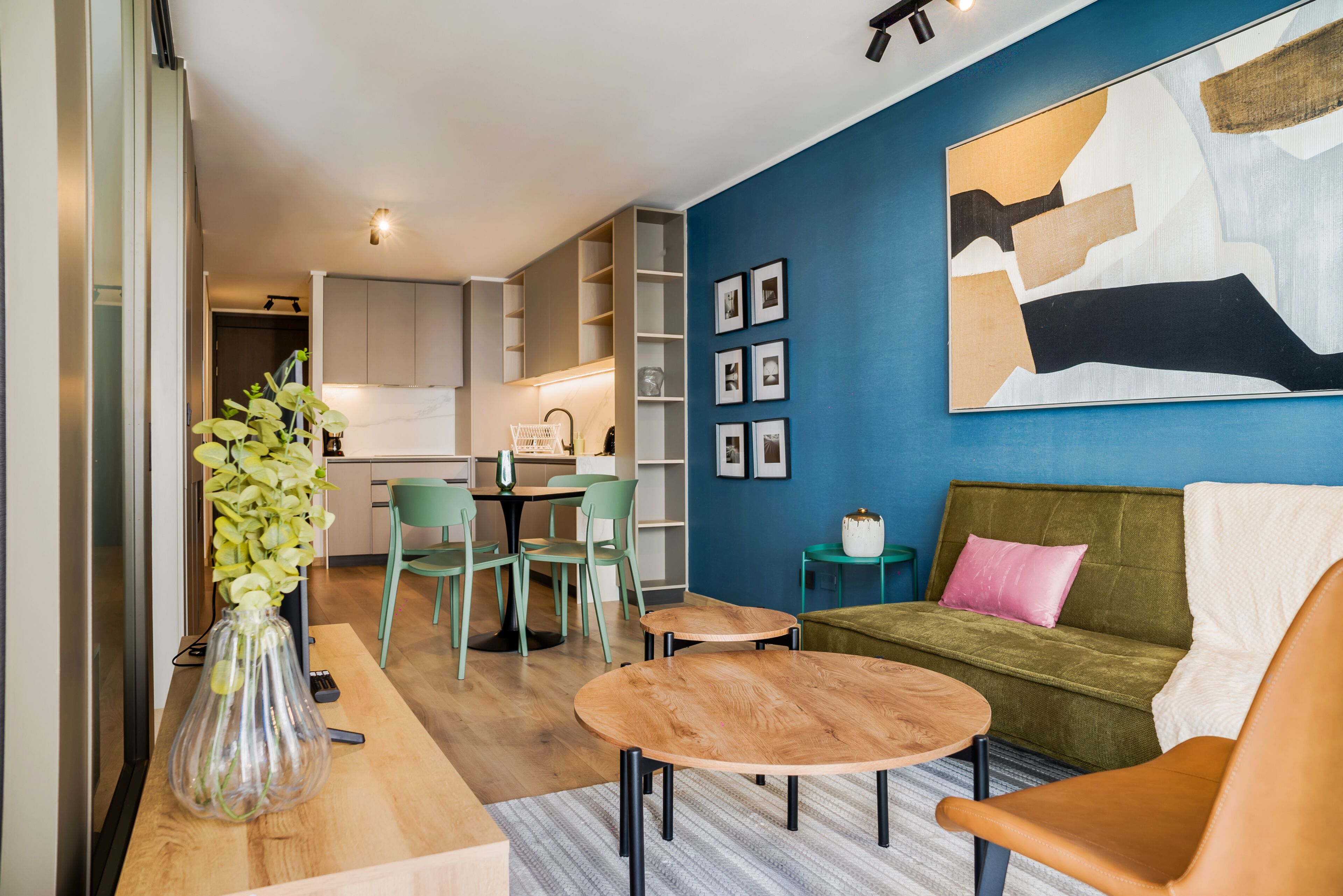 A modern living room with a sofa, coffee table, and chairs. There is a dining area with a table and chairs. The room has a blue accent wall and a large abstract painting.