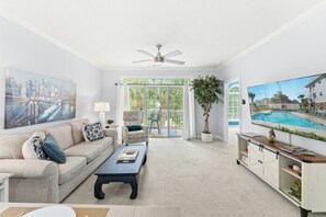 A living room with a sofa, coffee table, TV, and a large window with a view of a pool. - Barefoot Cypress 523- 2 Bedroom Condo (North Myrtle Beach)