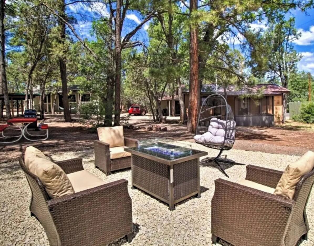 An outdoor seating area with wicker furniture and a glass-top table, surrounded by trees and a building in the background.