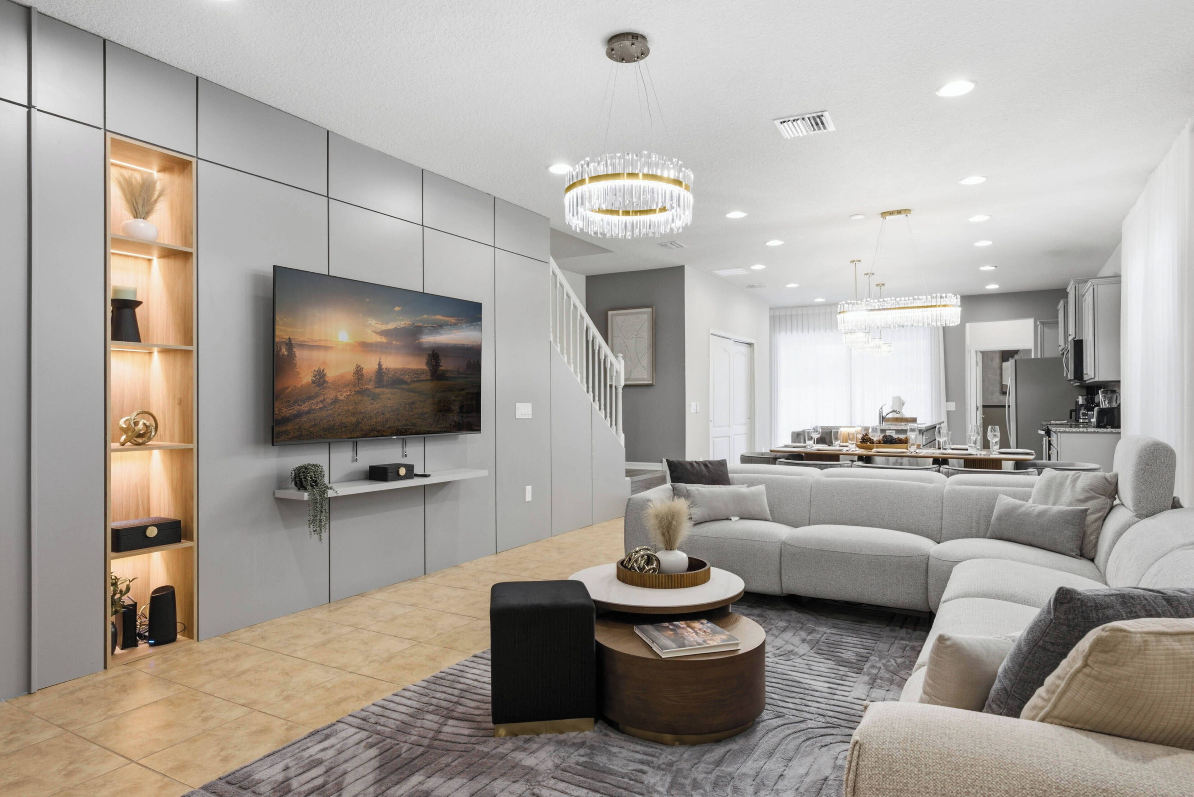 A modern living room with a large flat-screen TV, a sectional sofa, a coffee table, and a chandelier.