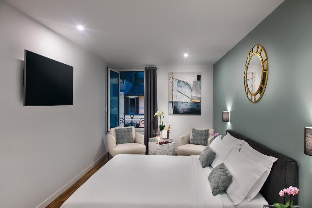 A modern hotel room with a large bed, two armchairs, a small table with a vase and flowers, a painting on the wall, and a mounted clock.