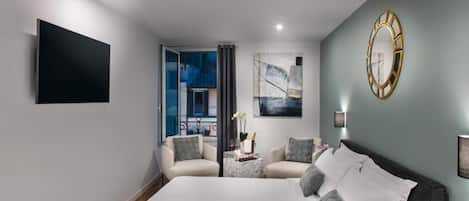 A modern hotel room with a large bed, two armchairs, a small table with a vase and flowers, a painting on the wall, and a mounted clock.