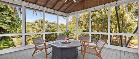 A covered outdoor patio with a fire pit table and chairs, surrounded by large windows and a wooden ceiling.