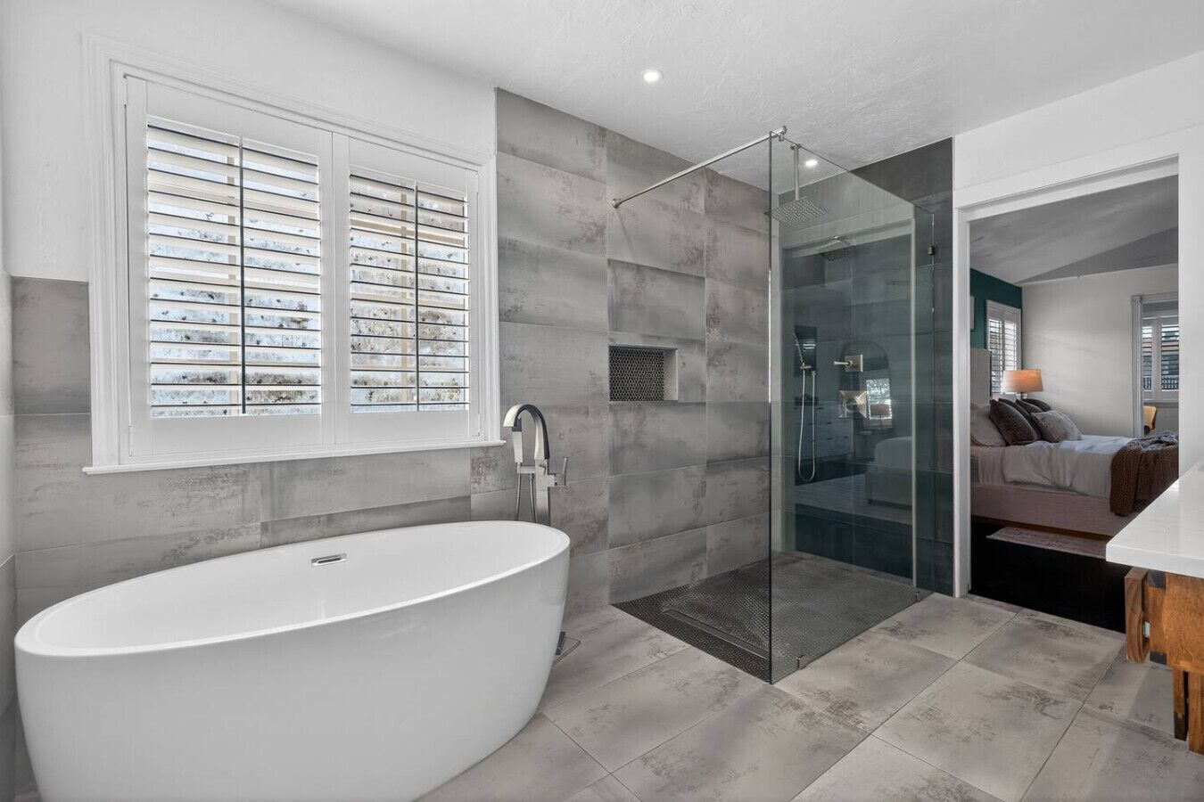 Un baño moderno con una bañera independiente, ducha con mampara de vidrio y ventana con persianas.