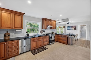A modern kitchen with wooden cabinets, stainless steel appliances, and a large window providing natural light. - Elite Living | Kayak | Joy Pool | Outdoor Kitchen ! (Fort Myers)