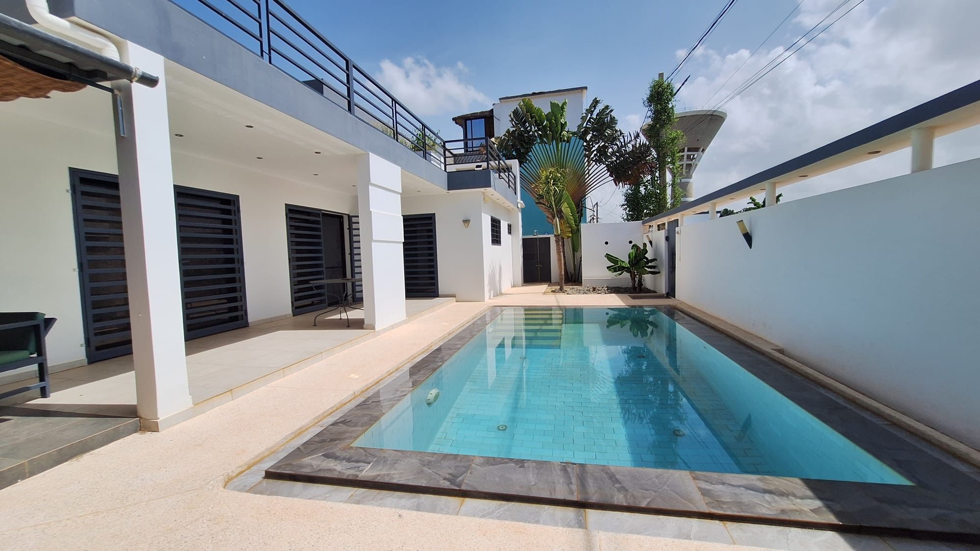 A modern residential pool with a clear blue water surface, surrounded by a white exterior wall, a covered patio area, and a small garden with palm trees.