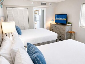 A hotel room with two beds, a television, a desk with a lamp, and a view of the kitchen. - Blue Haven by StayMareni - Oceanfront Suite (Myrtle Beach)
