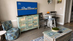 A living room with a television, a blue sofa, a coffee table, and a dining area with chairs and a table. - Blue Haven by StayMareni - Oceanfront Suite (Myrtle Beach)