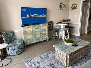 A living room with a television, a blue sofa, a coffee table, and a dining area with chairs and a table. - Blue Haven by StayMareni - Oceanfront Suite (Myrtle Beach)