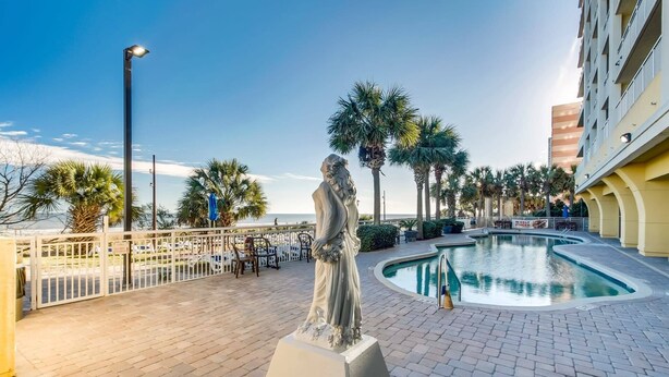 A statue in a courtyard with a pool, palm trees, and a building in the background. - Blue Haven by StayMareni - Oceanfront Suite (Myrtle Beach)