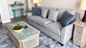 A living room with a grey sofa, two side tables, a coffee table, and a wall-mounted shelf. - Blue Haven by StayMareni - Oceanfront Suite (Myrtle Beach)