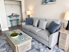 A living room with a grey sofa, two side tables, a coffee table, and a wall-mounted shelf. - Blue Haven by StayMareni - Oceanfront Suite (Myrtle Beach)