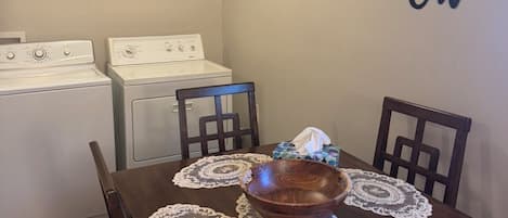Un comedor con una mesa de madera, individuales de encaje, un cuenco de madera y una lata de café. Se ven dos electrodomésticos blancos al fondo, y una pared con un letrero que dice 'Let's Eat'.