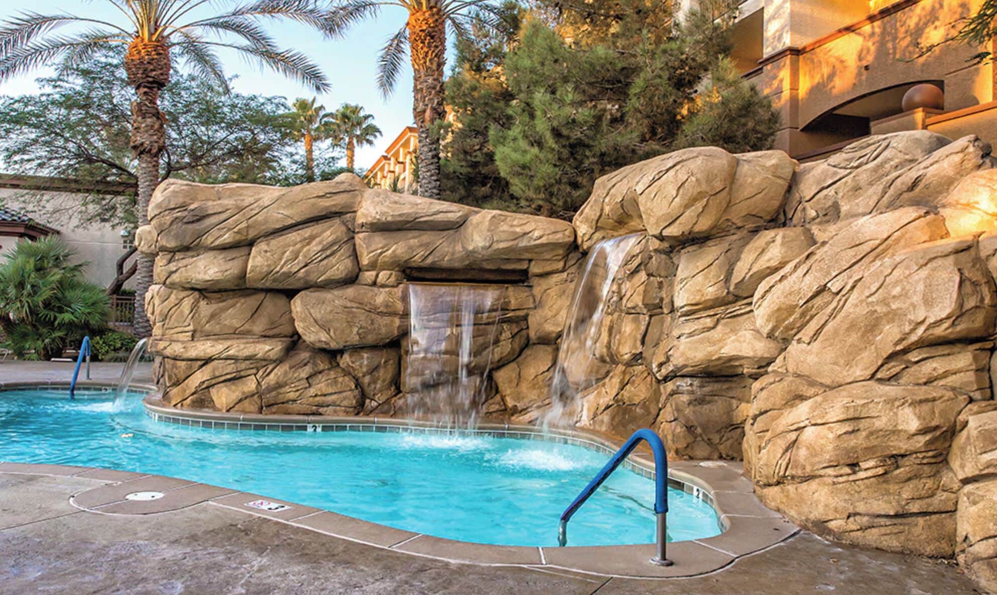 Una piscina con una cascada, rodeada de grandes rocas y palmeras.