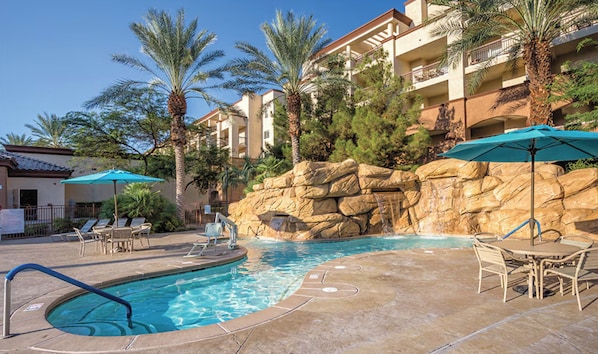 A swimming pool with lounge chairs and umbrellas, surrounded by palm trees and a rocky wall.