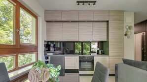 Uma cozinha moderna com eletrodomésticos embutidos, um espaço de refeições com cadeiras estofadas em cinza e uma grande janela com vista para árvores.