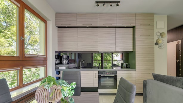 A modern kitchen with built-in appliances, a dining area with grey upholstered chairs, and a large window offering a view of trees. - Sopot Aquarius Bay by Lion Apartments (Sopot)