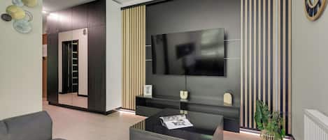 A modern living room with a flat-screen TV mounted on a dark wall, a coffee table with magazines, and a grey sofa.