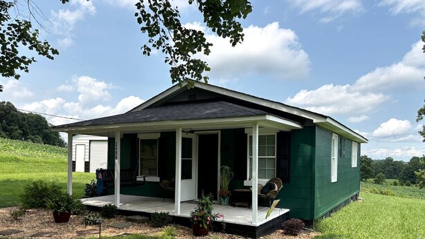 Eenlaags huis met een veranda, omgeven door een grasveld en bomen.
