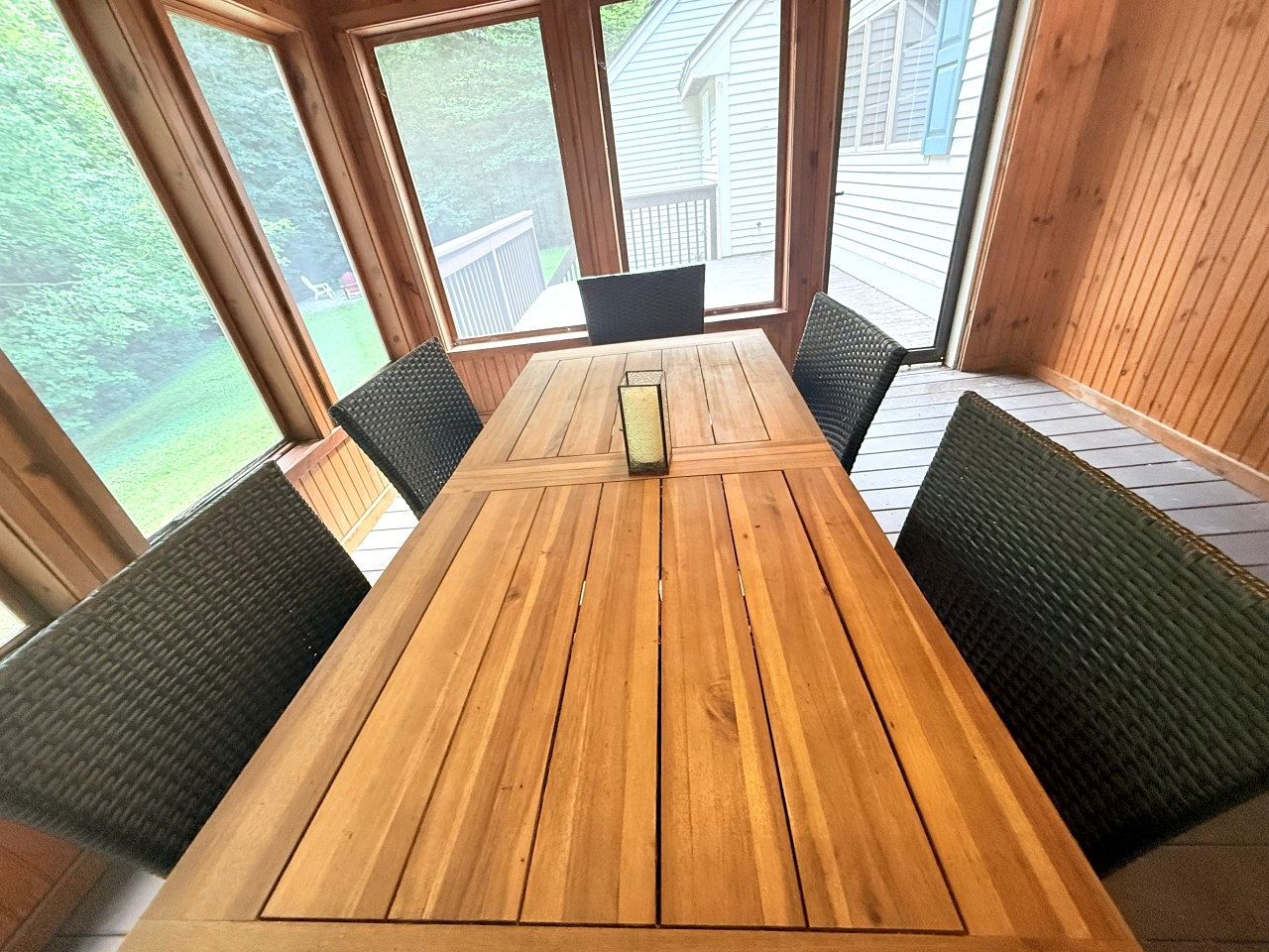 A wooden dining table with four chairs, large windows, and a view of a deck and greenery.
