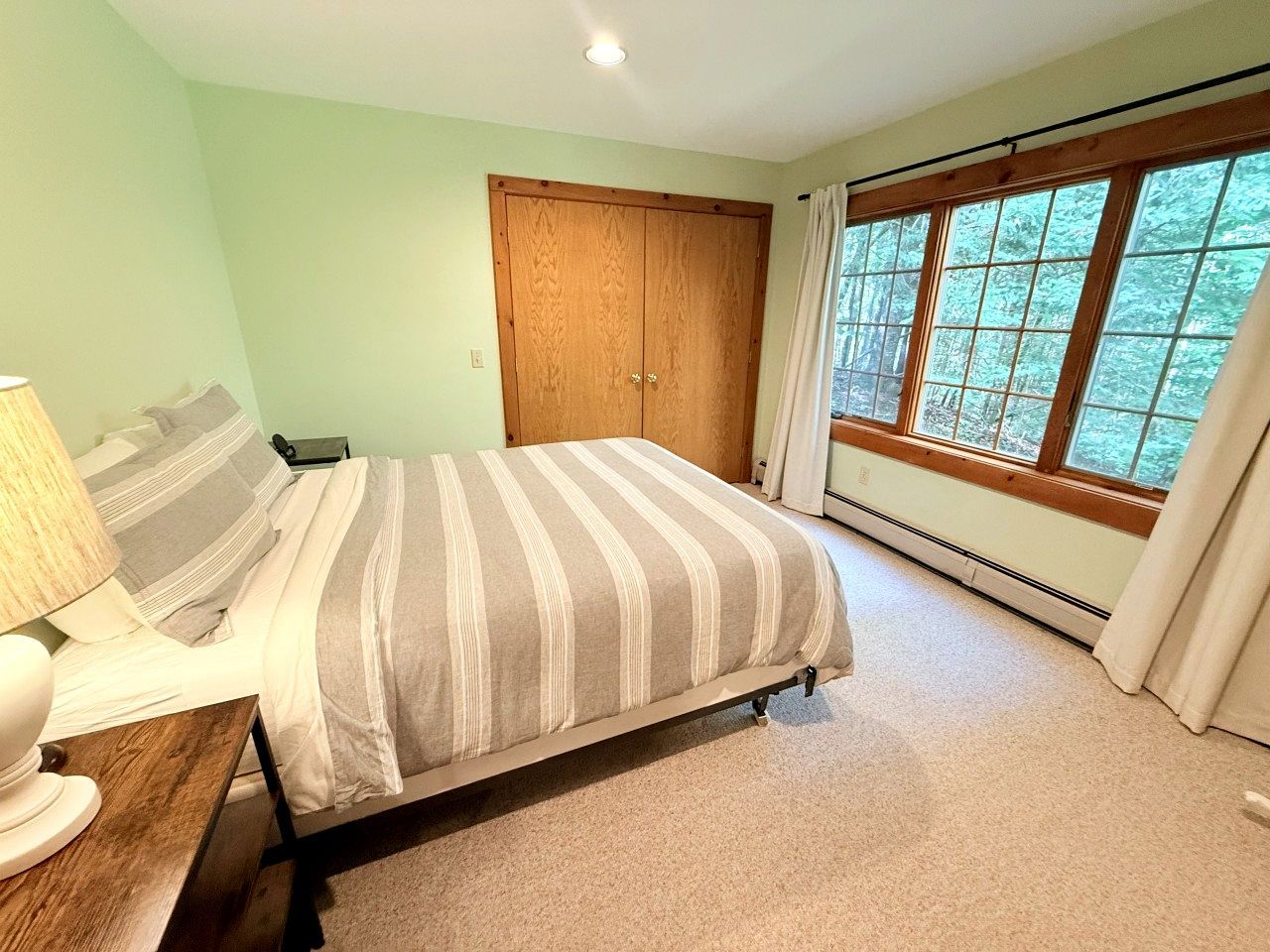 Een slaapkamer met een bed, een nachtkastje, een lamp, een raam met gordijnen en een houten garderobe.