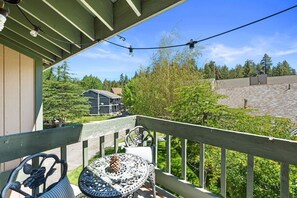A wooden balcony with a table and chairs, overlooking a house and a paved area with trees. - Lakeside Vibes | 2BR, Walk to Boulder Bay, Pool (Big Bear Lake)