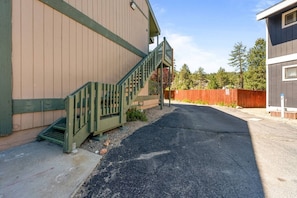 A building with a green staircase leading to a door, a paved driveway, and a wooden fence in the background. - Lakeside Vibes | 2BR, Walk to Boulder Bay, Pool (Big Bear Lake)