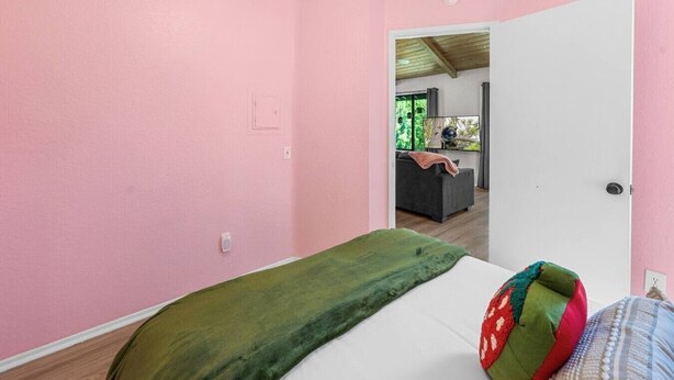 A room with pink walls, a bed with a green blanket, and a door leading to another room with a sofa and a window. - Lakeside Vibes | 2BR, Walk to Boulder Bay, Pool (Big Bear Lake)