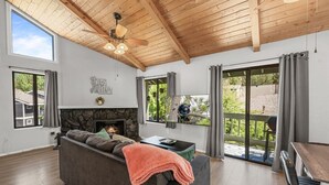A modern living room with a fireplace, a sofa, a dining table, and a large window with a view of greenery. - Lakeside Vibes | 2BR, Walk to Boulder Bay, Pool (Big Bear Lake)