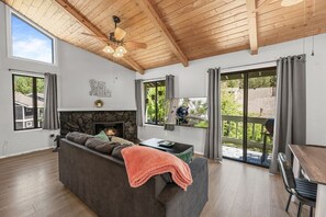 A modern living room with a fireplace, a sofa, a dining table, and a large window with a view of greenery. - Lakeside Vibes | 2BR, Walk to Boulder Bay, Pool (Big Bear Lake)