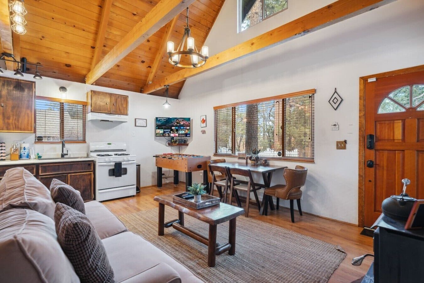 A cozy living room with a sofa, a coffee table, and a dining area with chairs and a table. There is a kitchen with a stove and a foosball table.