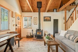 Ein gemütliches Wohnzimmer mit Kamin, Holzbalken, einer Couch, einem Tisch und einem Fenster mit Jalousien.