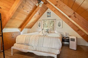 Une chambre avec un plafond en pente, un lit recouvert d’une couvre-lit à motifs, une table de chevet, une lampe et un climatiseur fixé au mur.